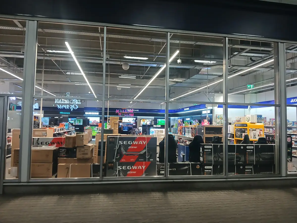 Shop window at Media Galaxy Craiova with boxed Segway electric scooters, gaming chairs and stacked stock, seen from the walkway at night, September 2026