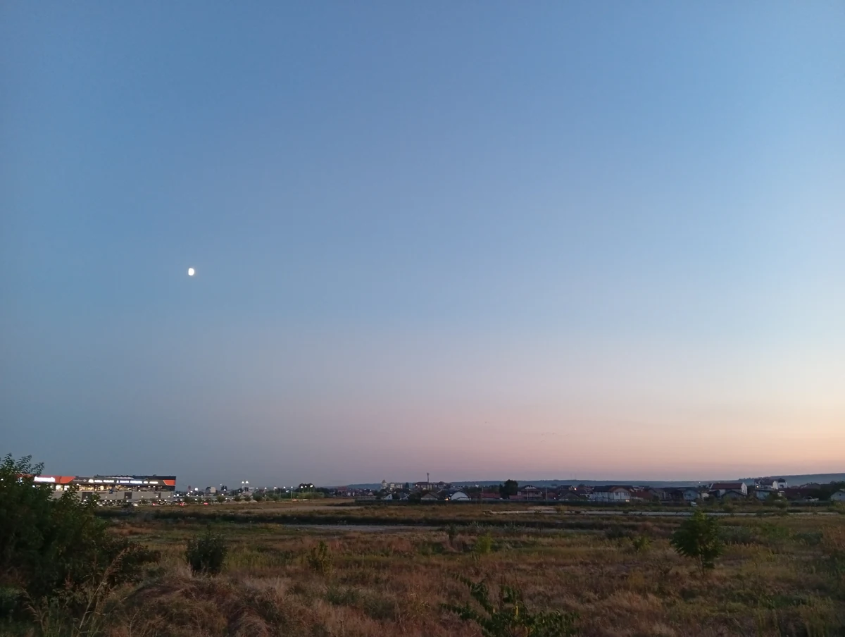 The moon over open ground on the western edge of Craiova at dusk, a lit shopping centre and houses on the horizon, 20 September 2026