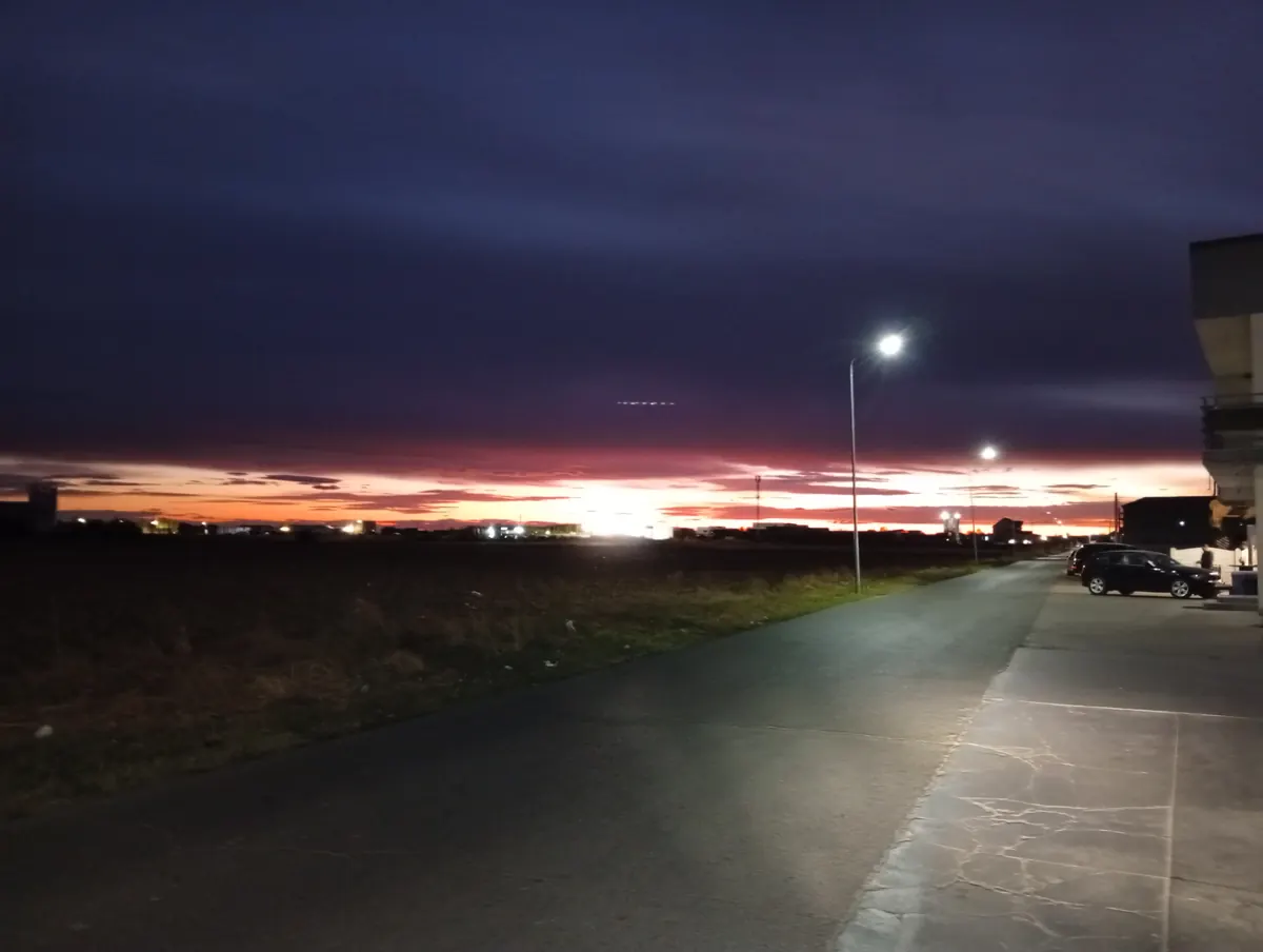 Red sunset over a ploughed field about two kilometres south of Craiova airport, a lit street and parked cars on the right and an aircraft's lights crossing the cloud band, 23 September 2026