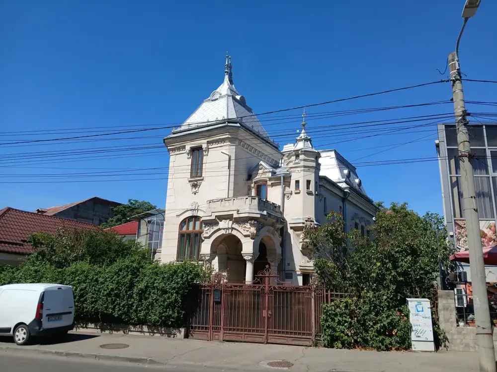 Number 31, not yet identified: a villa of about 1900 with a zinc-scaled pyramid roof and spire, a corner turret, and a double-arched loggia on squat columns under a stone balcony, all of it behind a rusting Art Nouveau gate. A heritage plaque is fixed to the pier by the door, which is how we know it is listed and not what it is called. If you can read the plaque, tell us