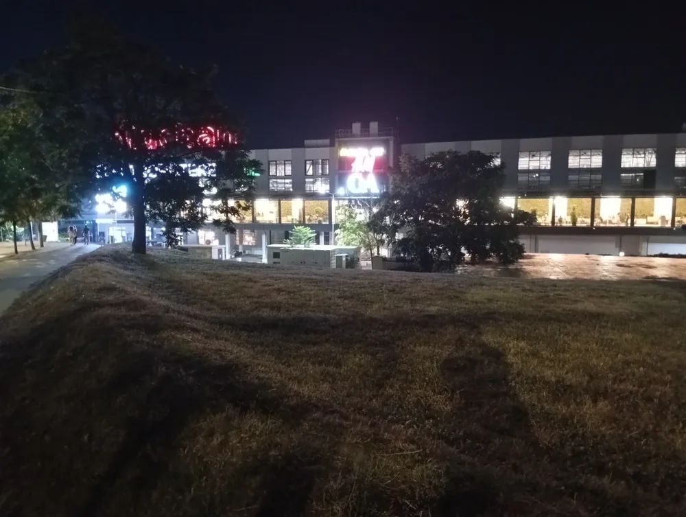 The whole block from across the grass — Auchan in red neon, H&M and C&A lit above the entrance, the glazed gallery running the length of the building