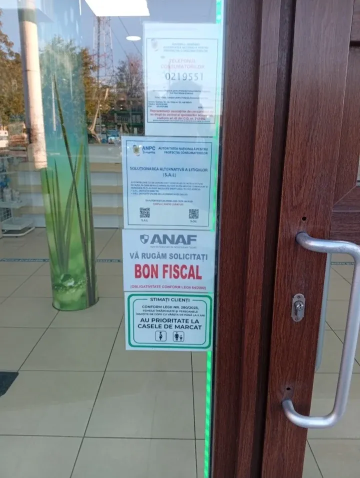 The standard stack, stuck to a glass door in Craiova: the ANPC hotline, the SAL/SOL dispute notice, the ANAF receipt reminder, and the priority-till notice